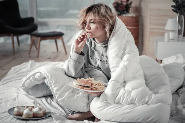 Woman wrapped up in a blanket eating snacks and giving into food cravings